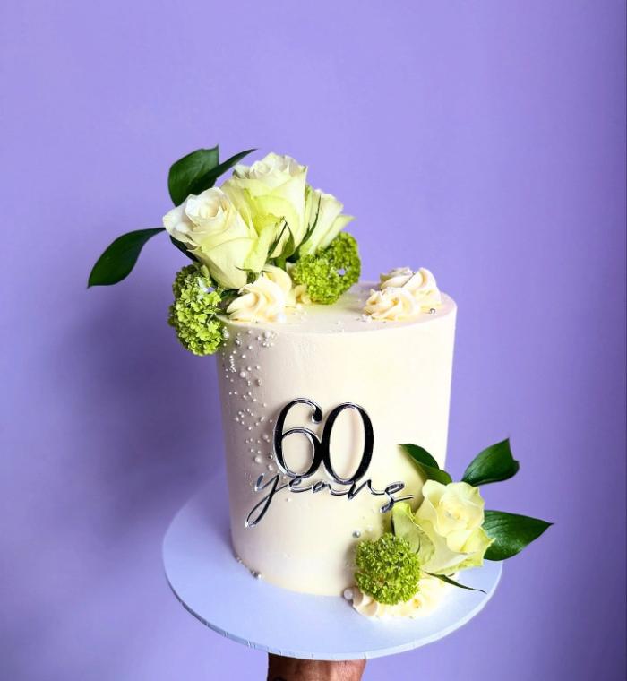 A white buttercream cake dressed with fresh white roses and greenery, topped with a sixty years topper