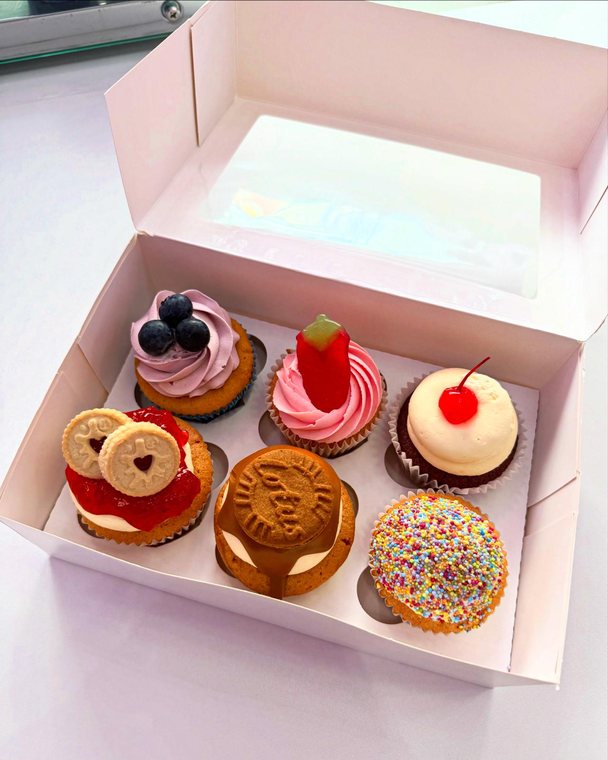 An open pink box of six cupcakes, each topped differently with blueberries, strawberry, cherry, a jammie dodger, Biscoff and sprinkles