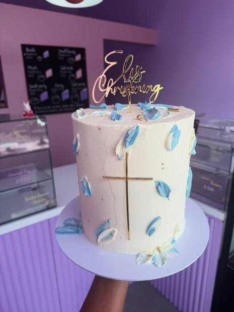 A tall white buttercream christening cake with a gold cross, gold script topper and pale blue feather detail