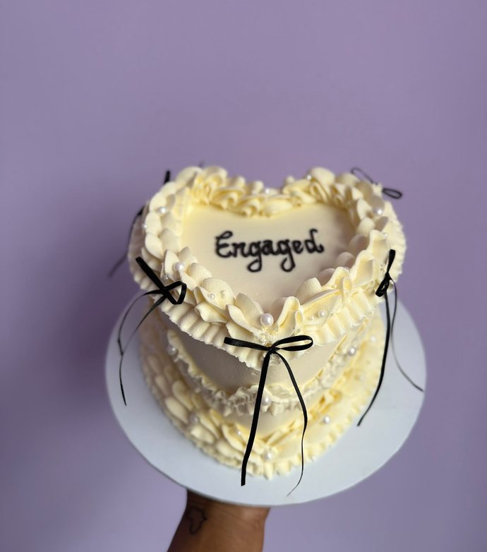 A cream buttercream heart shaped cake with ruffled piping, pearls, black bows and the word Engaged