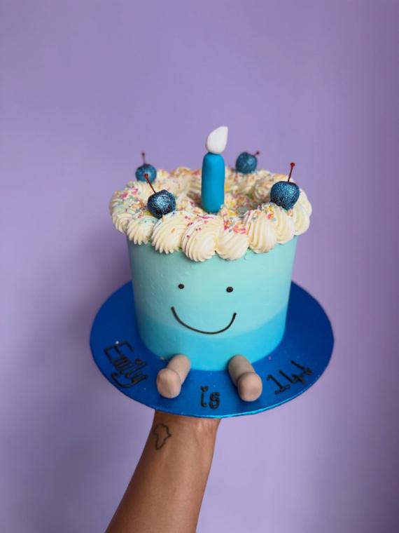 A pale blue buttercream cake with a smiling face, piped swirls and a candle, made for a small child's birthday