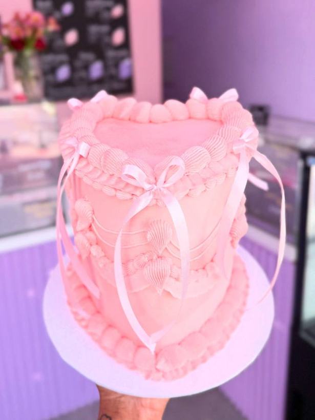 A tall pink buttercream heart cake finished with a piped ruffle edge and a ribbon bow